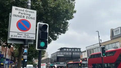 A parking restrictions sign on Wood Green High Street, with traffic lights, red buses and buildings in the background