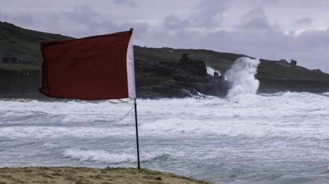 Waves crash up over the rocks and a red flag blowing in the foreground