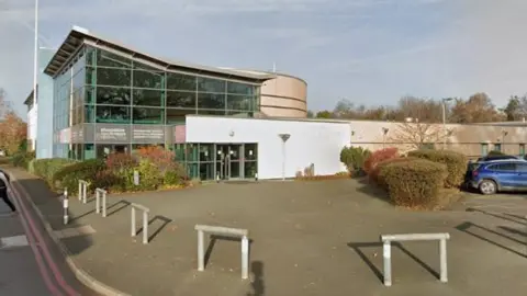 A large building with a glass wall and white entryway. There is a car park on the right hand side