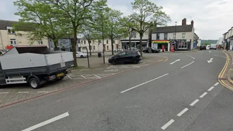 A Google Street view image from a junction showing a car park on the left with trees and two vehicles parked up. A road curves round to the left behind the car park. High street shops line the side of the street.