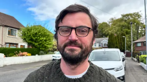 A man with curly brown hair and a beard. Wearing black rimmed glasses and a green knitted jumper. He is staring into camera with neutral expression. He is stood on a residential street in front of a white car.