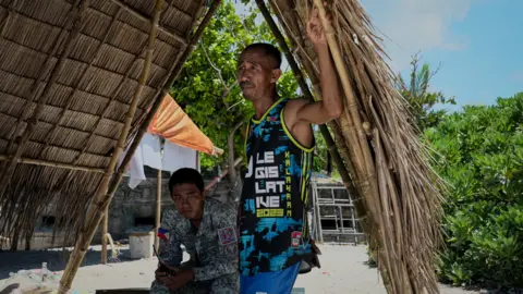 Two men stand in the shade of a shelter made of reeds
