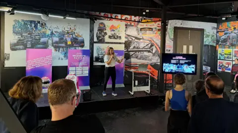 Silverstone Museum Racing driver Caitlin Wood is standing in front of racing car artwork and holding a microphone. In front of her is an audience sitting on chairs listening to her.