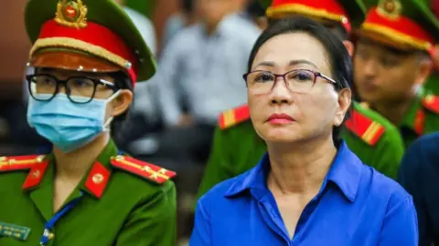 Truong My Lan wearing glasses and a blue shirt, sitting beside an official in uniform.