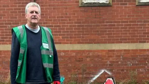 Crewe Clean Team A man with short grey hair, wearing a green jacket and dark jumper, stands in front of a building's brick wall.