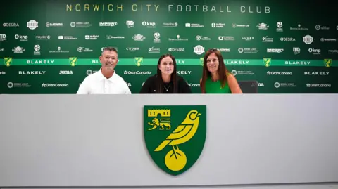 Supplied Eloise King sits between her parents, on the press conference desk. The desk is grey, and has a big Norwich City emblem. 