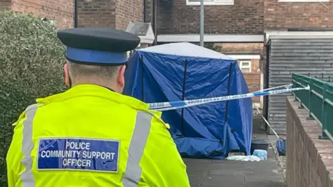 A police community support officer stands in front of a blue forensic tent.