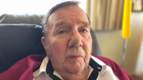 BBC Russell sat in an armchair. He has short grey hair and has a moustache.