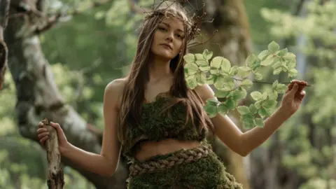 Stock of a mythical woman in the forest. She has long brown straight hair and is holding a branch in one and a stick in the other