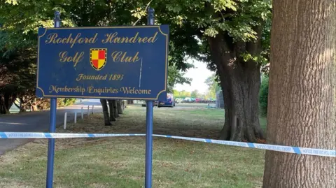 Nick Hull/BBC A blue sign that reads Rochford Hundred Golf Club, with police crime scene tape beneath it block the driveway to the clubhouse. There are a number of trees either side of the entrance.