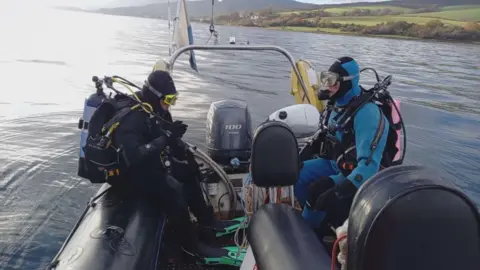 Howard Wood/Coast Two men in full diving gear, including oxygen tanks, sit on a rigid inflatable dingy ready to fall into the water