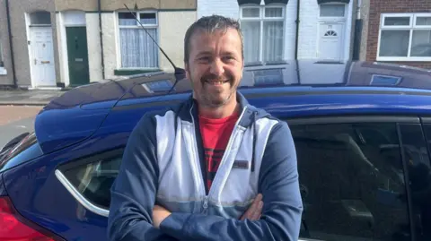 Scott Holliday stands outside a car with his arms folded. He is smiling. He has short grey hair and is wearing a grey and white hoodie.