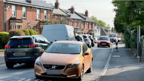 Church Road in Cheltenham during daytime. It is sunny. There is terraced housing down one side of the street. There is queuing stationary traffic heading in one direction and several cars travelling on the opposite side.