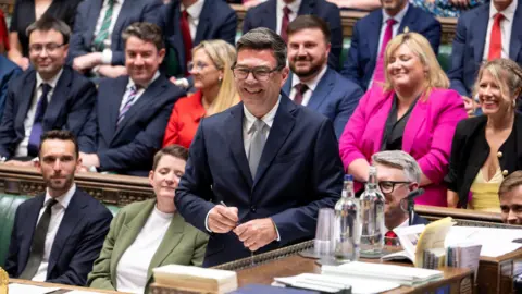 House of Commons Andy Burnham is pictured smiling in the House of Commons as he is sworn in as the MP for Makerfield. MPs can be seen in the background on the famous green benches while he holds a pen. He is wearing a dark suit jacket, grey tie and white shit. He wears glasses and has short, dark hair.