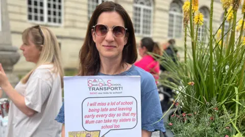 A woman wearing a blue top and dark glasses holds a sign saying 'Save Our School Transport'.