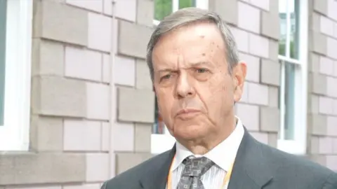 Gary Clueit, a man with short grey hair, wearing a white shirt, grey blazer and grey tie. He is standing in front of a pink granite building with white windows.
