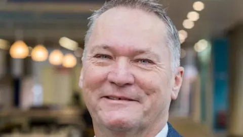 Michael Stratton is smiling at the camera in a close-up shot of his face. He has short, greying hair and is wearing a white shirt and blue jacket.