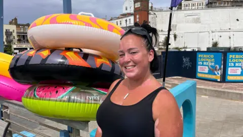 Phil Harrison/BBC A woman wearing a black top and sunglasses resting on her head. She is standing next to some inflatable rings.