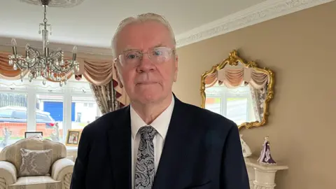 A man with a dark blue suit jacket and white shirt, with a Paisley-patterned blue and white tie looks forward towards the camera.
He is wearing glasses and has platinum coloured hair.
In the background is a clear chandelier, a puffy cream-coloured living room chair, a white mantlepiece with a doll in a purple dress on it.
There are tan walls and a large wall-mounted mirror with an ornate gold-coloured frame