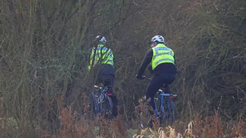 Peterborough Walks Two police officers, wearing green high-visibility jackets and white helmets, are cycling on a green and leafy cycle path.