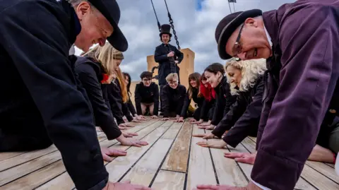 SS Great Britain Trust An actor dressed as Isambard Kingdom Brunel wearing a top hat and black dress attire with a waistcoat looks on as 10 people kneel with their hands flat on the deck of the SS Great Britain. 