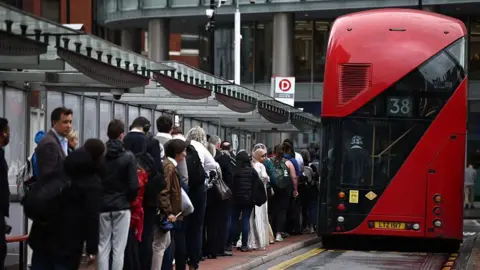 Getty Images A queue of people at a bus stop waiting to board a red London bus with the number 38 visible on the back of the bus.