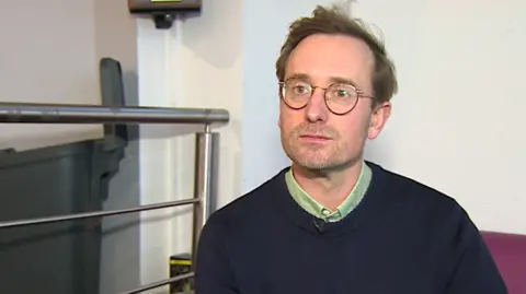 A man with short brown hair and round glasses. He is wearing a dark blue jumper and is sat on a sofa next to some metal railings.