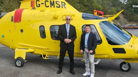 Stoke Air Ambulance Phil Copeland and Daniel Moores from toke air Ambulance are standing by a yellow helicopter. They vehicle is parked on a hard surface, with trees and fencing in the distance.