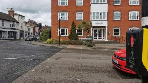 Ruth Leach A traffic light crossing. Across the road is a residential home that is three storeys tall. There is a red car waiting behind the traffic lights.