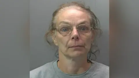 Cambridgeshire Police Custody photo of Melanie Wright. She has curly greying hair that has been tied back from her face and is wearing thin framed gold rimmed glasses. She is also wearing a plain grey sweatshirt.