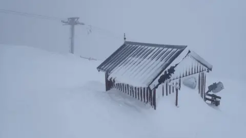 The hut has a corrugated roof and walls. It is almost buried by snow. A ski life is visible in the gloom behind it.