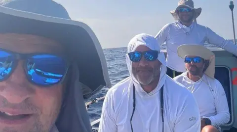 The Mayflower Atlantic Challenge Four rowers in hats and white tops and wearing sunglasses in a boat with sea behind.