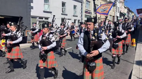 A pipe band walking along the road with red and green kilts. They are playing bagpipes and a banner can be seen flying in their midst.