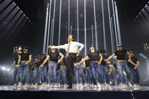 Getty Images Harry Styles dances on stage at the Brit Awards. Wearing a shirt, tie and pinstripe trouser combination, he leads a group of dancers dressed in jeans and black t-shirts.