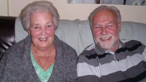 Mary with husband Ron. Ron died in 2016. They are both sat on a sofa smiling. 