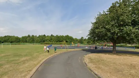 Google An open green space under blue skies. There is a set of goalposts and a path leads to a small playground where children are playing on the swings. Two people stand in the foreground talking. Trees are visible in the distance and on a smaller patch of grass near the playground.