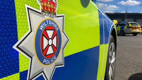 BBC Generic close-up shot of the side of a Wiltshire Police car showing the logo. Another police car can be seen in the background.