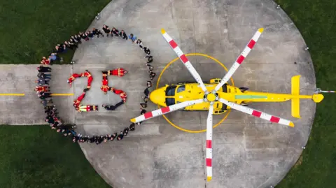 DSAA Aerial view of people standing in a circle around eight other people making the figure 25, next to a yellow helicopter.