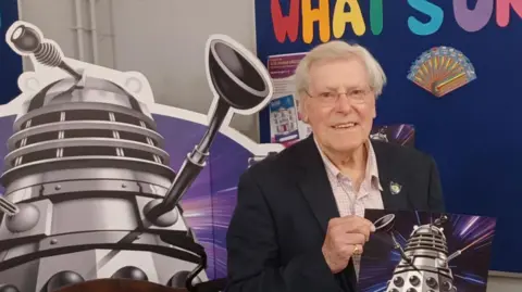 Paul Hayes/BBC Peter Purves, a white-haired man in his eighties, wearing glasses, a dark blue jacket, white checked shirt and light brown trousers, stands behind a desk, holding up a poster reading ‘Adventures in Space and Time’ and displaying an image of a Dalek, a conical robotic creature. Behind him is a large cardboard cut-out of a Dalek.