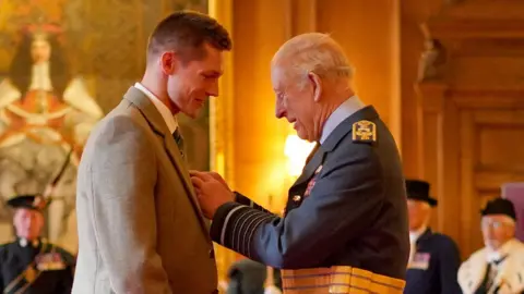 PA Media Swimmer Duncan Scott wearing a beige jacket and tie receives an OBE from King Charles who is wearing a dark-coloured jacket.