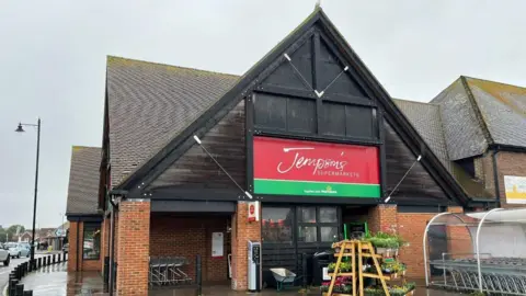 Jempson's Supermarket in Rye Town Centre has a green and red sign on the front and timber on the building and there are racks of plants outside with supermarket trolleys.