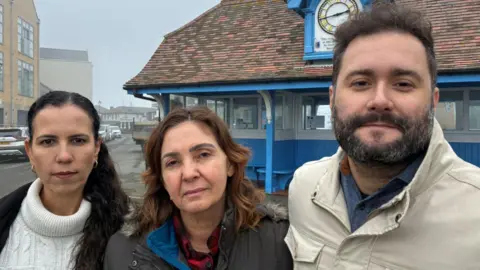 Two women and a man standing in front of a shelter next to a road. The shelter is painted blue, with a tiled roof, which has a clock on it.