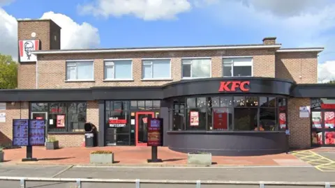 The exterior of a KFC restaurant in Broomfield Road, Chelmsford. It has a black trim frontage. The KFC branding is on the ground floor, but there is also an upper storey. There is blue sky and clouds above.
