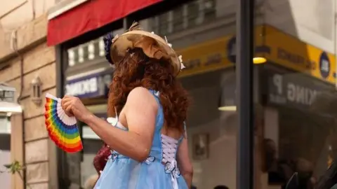 Getty Images A drag performer is in a street. They're wearing a curly auburn wig and a blue dress, that is laced up at the back and holding a rainbow coloured fan.
Copyright