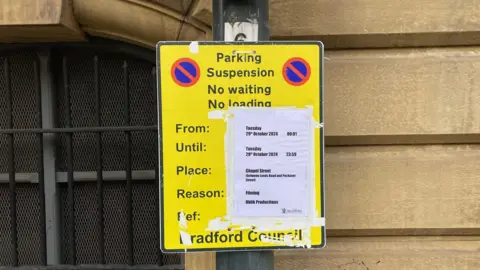 A yellow street sign on a lamppost with a white piece of paper taped over it showing the times when parking will not be available due to filming.  