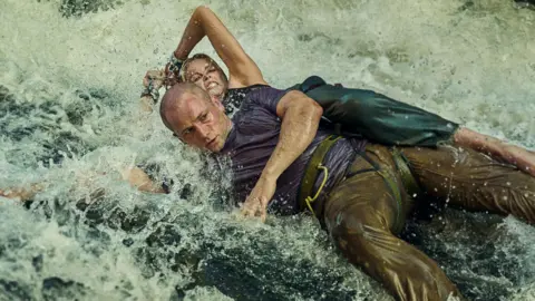 Egerton and Theron both cling to a large bolder in a river, with water crashing over them. Egerton wears a blue t-shirt and brown trousers, while Theron wears green trousers.