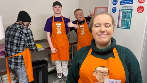 A woman in a teal rollneck jumper and an orange apron is standing in a working kitchen. Behind her are three men wearing the same apron, printed with a cartoon face of a young man wearing glasses. The man on the left has his back to camera, is wearing a black woolly hat and a checkered shirt, and is chopping using a yellow chopping board. Next to him is a man in a black baseball cap, purple t-shirt and white trousers holding his thumb up. The other man has short blonde hair and is wearing a black t-shirt. He is pointing to the sky in a pose. There is a black bin, red calendar and large white frideg covered in safety stickers. 