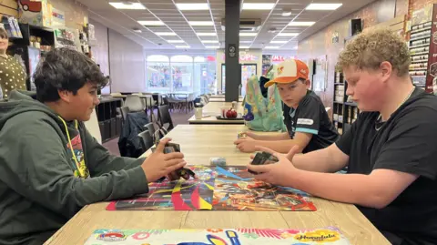 Darren Rozier/BBC The three boys sit in a cafe at a table playing Pokemon cards.
