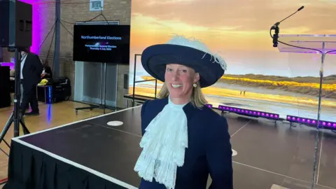 BBC Lucia Bridgeman smiling at the camera, wearing her navy blue hat with white feathers