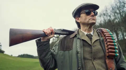 Disney+ David Tennant in costume wearing hunting gear and flat cap, and carrying a rifle and ammunition over his shoulder